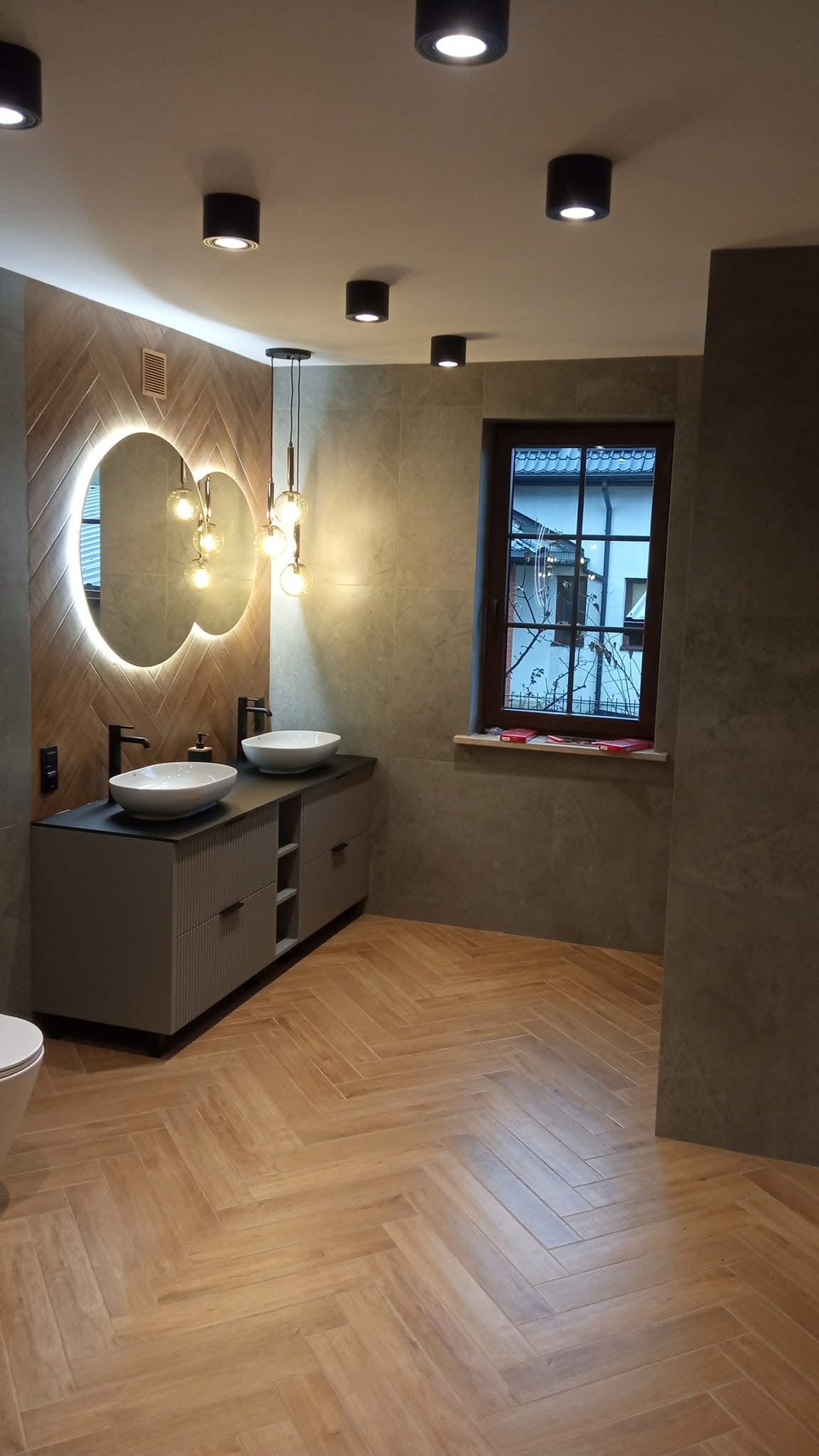 Elegancka łazienka po remoncie: drewnopodobna podłoga w jodełkę, podwójna umywalka nablatowa, okrągłe lustro z podświetleniem LED i nowoczesne oświetlenie sufitowe.