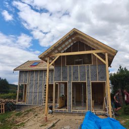 Drewniane Marzenia Damian Dyka - Konstrukcja domu szkieletowego w trakcie budowy. Widoczne drewniane ramy, izolacja Cry-X, fragment dachu z oknem. Na zewnątrz dwaj mężczyźni i niebieska plandeka.