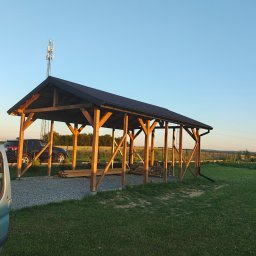 Drewniane Marzenia Damian Dyka - Drewniana wiata z czarnym dachem na żwirowym podłożu, widok z zewnątrz, częściowo widoczny samochód dostawczy, w tle krajobraz polny.