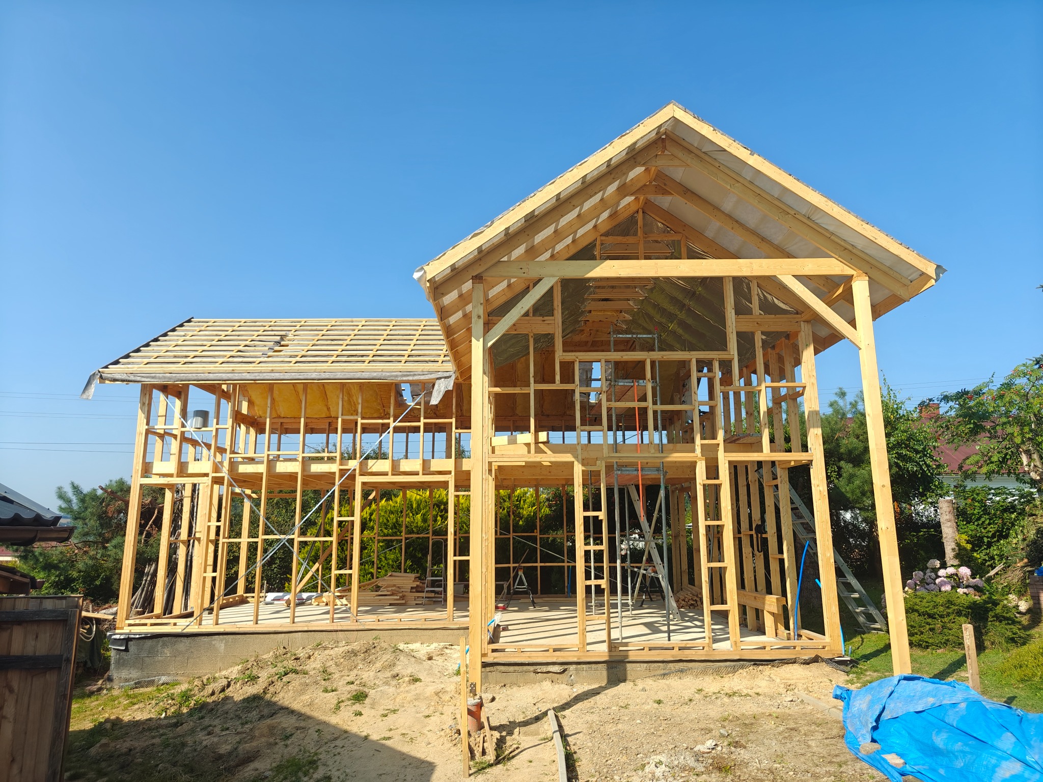 Surowy szkielet domu w trakcie budowy, z częściowo położonym dachem. Widoczne drewniane elementy konstrukcyjne i niebieska plandeka w tle.