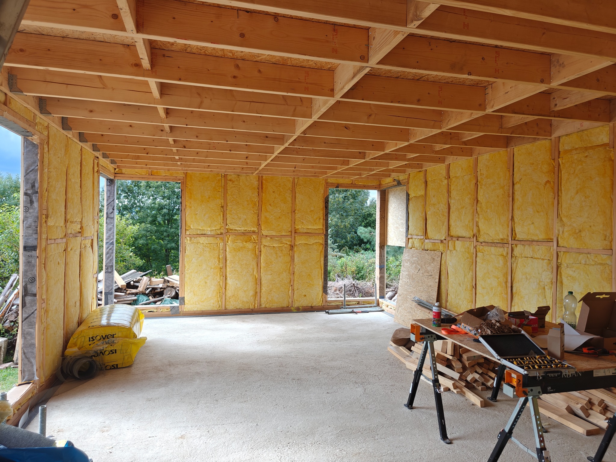 Wnętrze domu szkieletowego w trakcie budowy: drewniana konstrukcja ścian wypełniona izolacją, widoczne narzędzia i materiały budowlane. Surowy stan wykończenia.