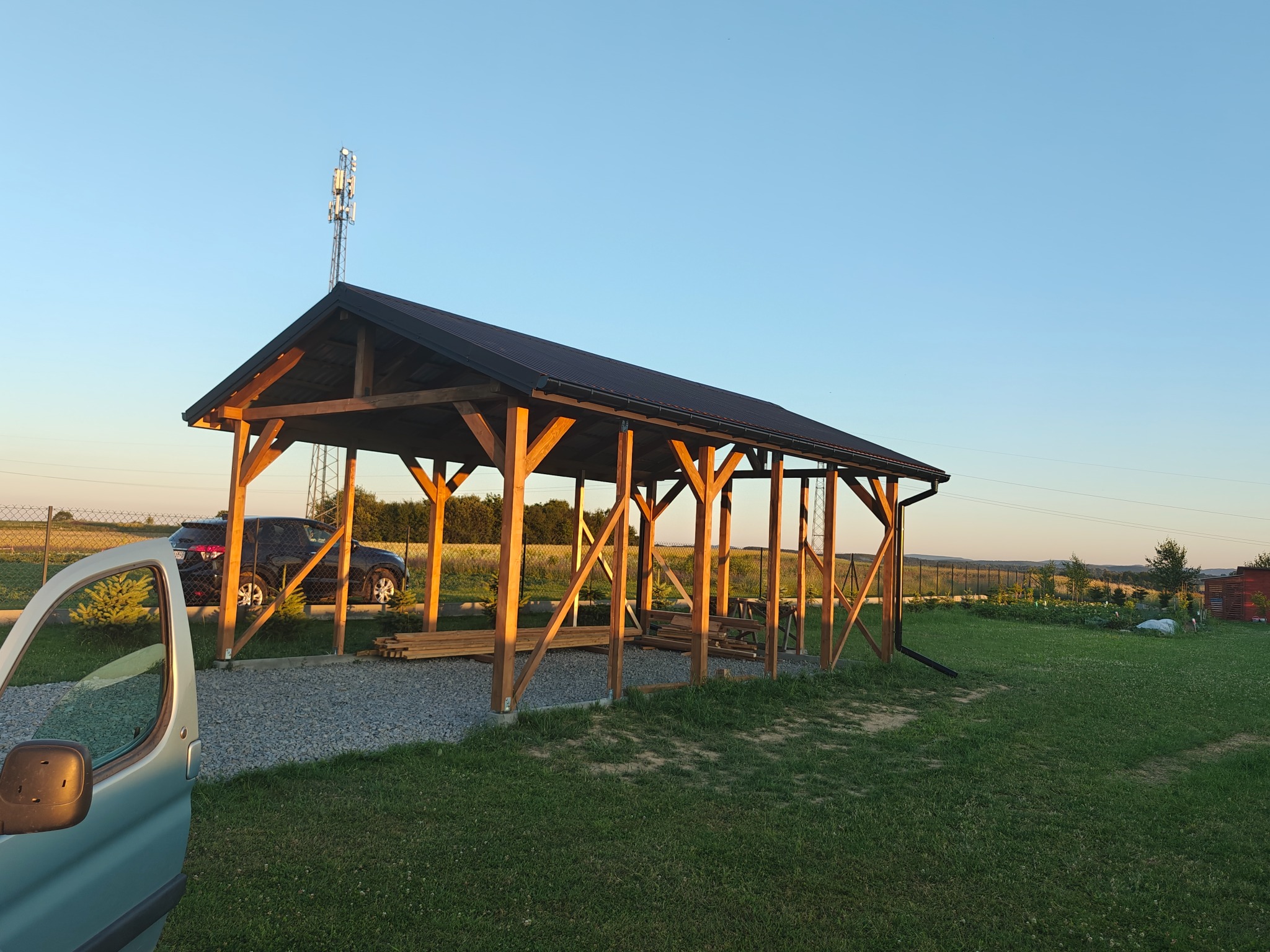 Drewniana wiata z czarnym dachem na żwirowym podłożu, widok z zewnątrz, częściowo widoczny samochód dostawczy, w tle krajobraz polny.