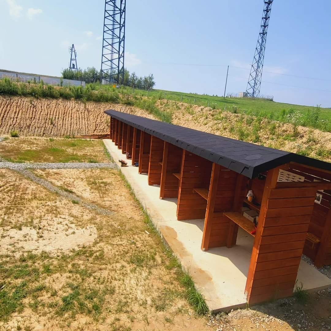 Długi, drewniany budynek z czarnym dachem na zewnątrz, widziany z perspektywy. W tle wzgórza i wysokie maszty telekomunikacyjne.