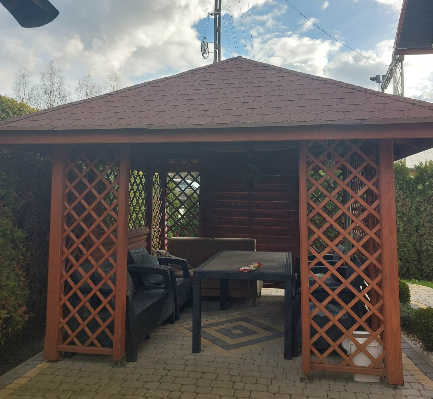 Drewniana altana ogrodowa z brązowym dachem z gontu bitumicznego, wyposażona w meble ogrodowe, na tle zieleni i błękitnego nieba z chmurami.