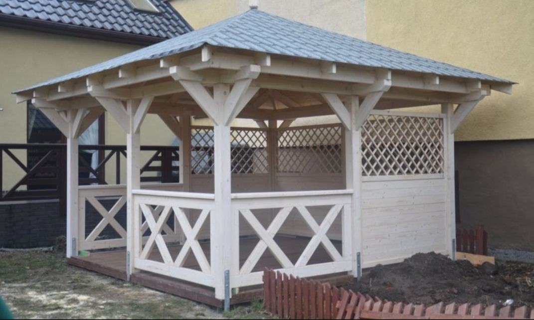 Drewniana altana ogrodowa z szarym dachem, białą konstrukcją i dekoracyjnymi kratami, prezentowana na tle budynku i fragmentu ogrodu.