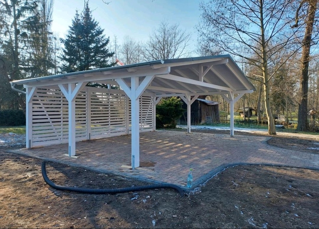 Biała, drewniana wiata garażowa z ażurowymi ścianami bocznymi, wyłożona kostką brukową, na tle drzew i błękitnego nieba.