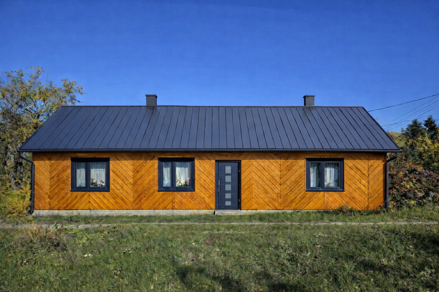 Parterowy dom z drewnianą elewacją ułożoną w jodełkę, kryty blachą na rąbek stojący, z ciemnymi oknami i drzwiami, na tle błękitnego nieba.