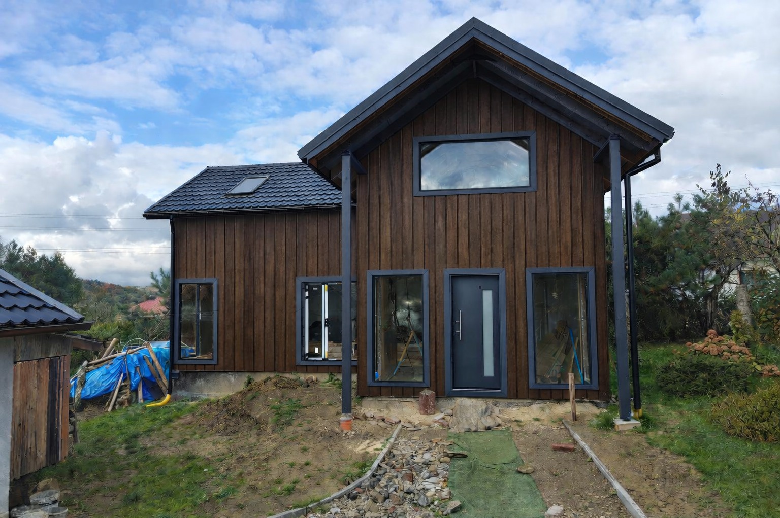 Surowy dom szkieletowy z ciemnego drewna we Frysztaku, z szarą stolarką okienną i drzwiami, w trakcie budowy, na tle zieleni i błękitnego nieba.