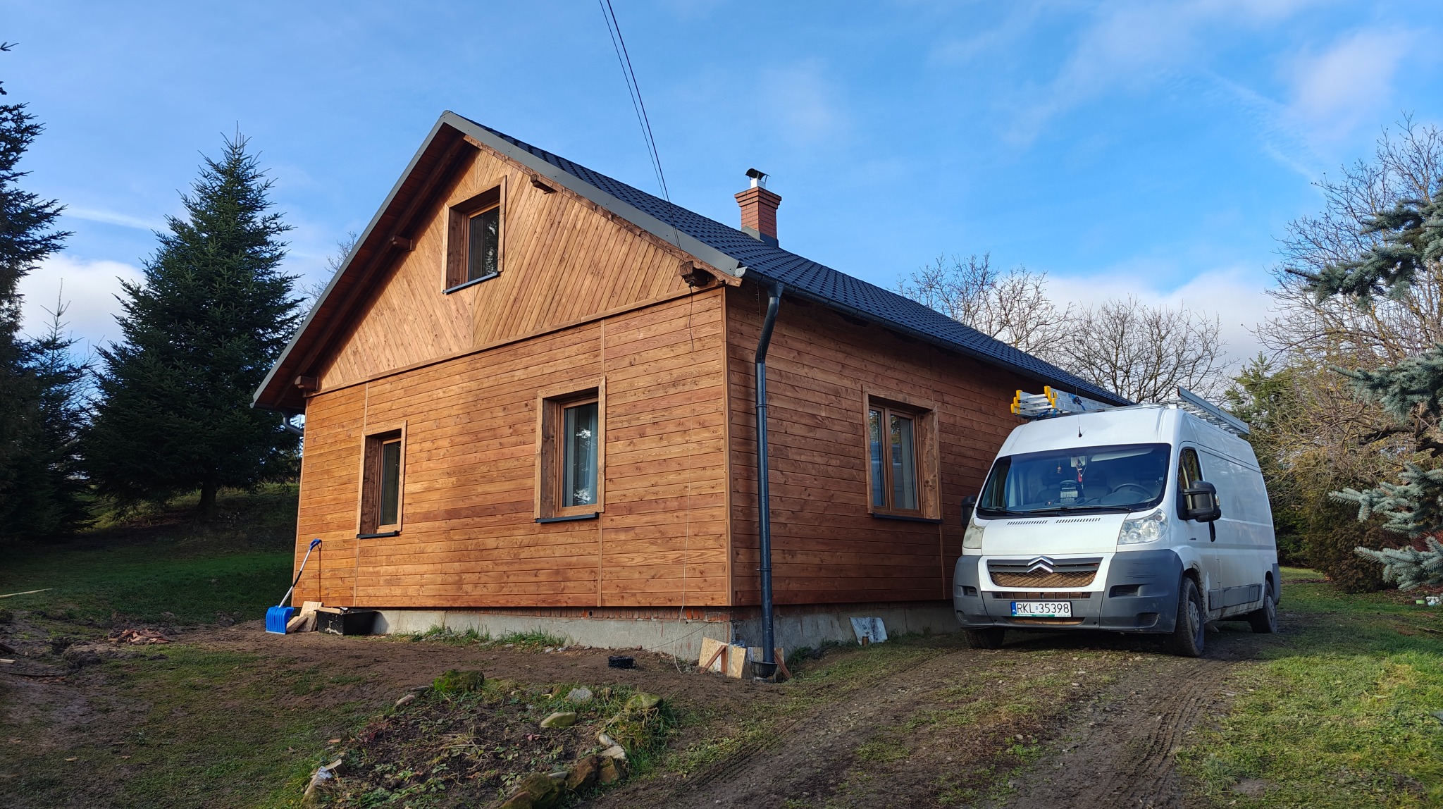 Nowoczesny dom szkieletowy z drewnianą elewacją, widoczny z boku, z zaparkowanym białym busem z drabiną na dachu. Błękitne niebo w tle.