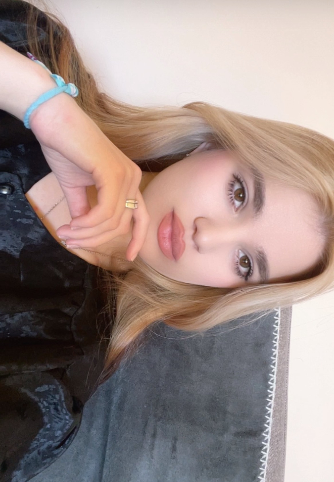 Portret młodej kobiety z blond włosami, delikatnym makijażem i złotym pierścionkiem, opierającej dłoń o szary materiał. Ujęcie z bliska, z boku.