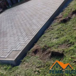 Jasnoszara kostka brukowa ułożona w jodełkę, obrzeża betonowe, trawa. Widok z góry, lekki kąt. Na trawie widoczny logotyp firmy.