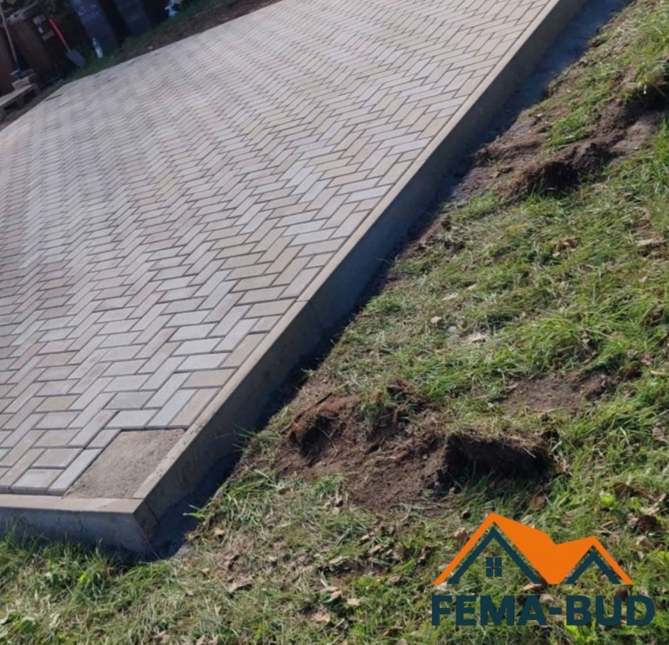 Jasnoszara kostka brukowa ułożona w jodełkę, obrzeża betonowe, trawa. Widok z góry, lekki kąt. Na trawie widoczny logotyp firmy.