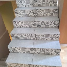 Ret Brother's Stanisław Retkowski - Schody wyłożone szarymi płytkami z dekoracyjnym wzorem patchworkowym na krawędziach stopni, widok z dołu, drewniana podłoga, jasne wnętrze.