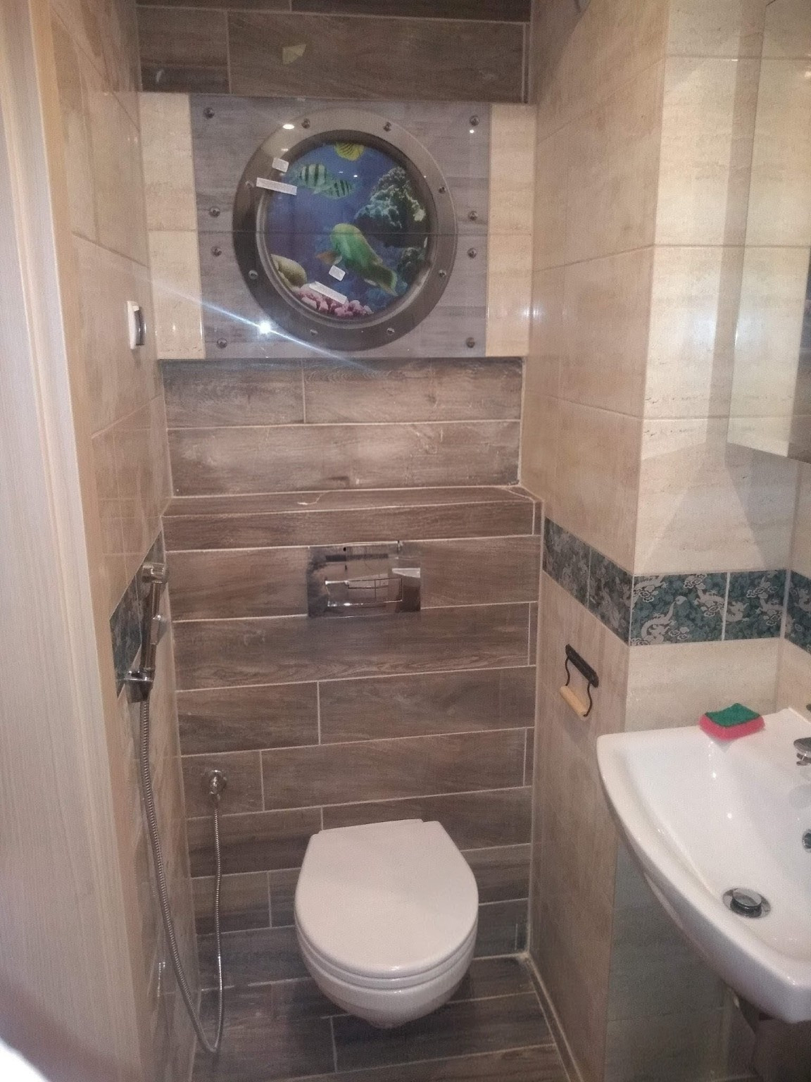 Wykończona toaleta z oryginalnym akcentem: okrągłe okno z motywem akwarium w płytkach. Umywalka z baterią i akcesoriami. Płytki imitujące drewno na ścianach i podłodze.
