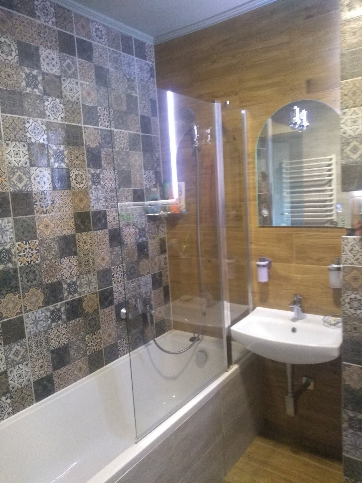 Wykończona łazienka z wanną za szklanym parawanem, umywalką na ścianie i lustrem w kształcie łuku. Ściany wyłożone mozaiką i panelami imitującymi drewno.