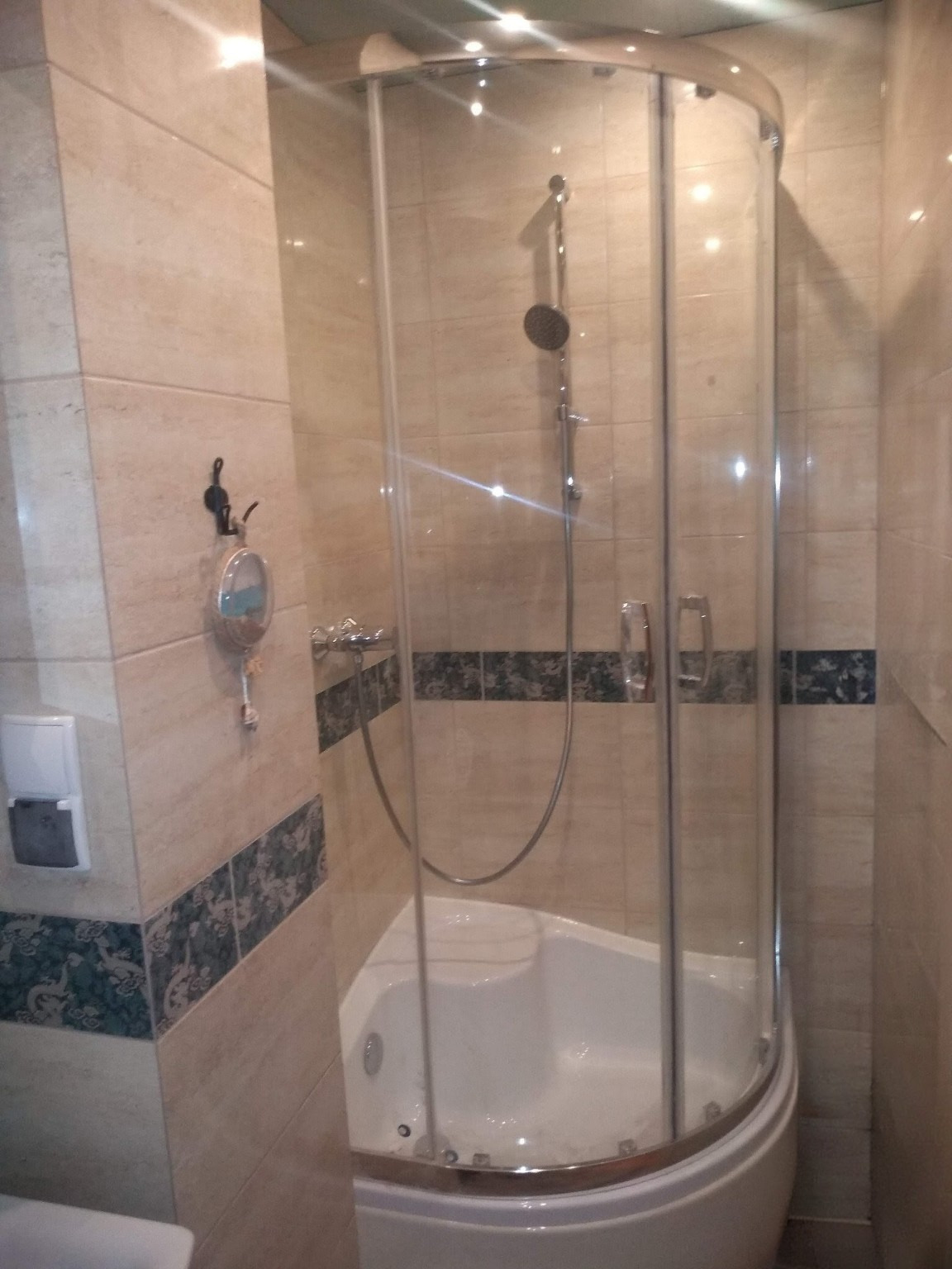 Narożna kabina prysznicowa z hydromasażem w jasnej łazience z beżowymi płytkami i dekoracyjnym pasem. Widoczny chromowany zestaw natryskowy i akcesoria łazienkowe.