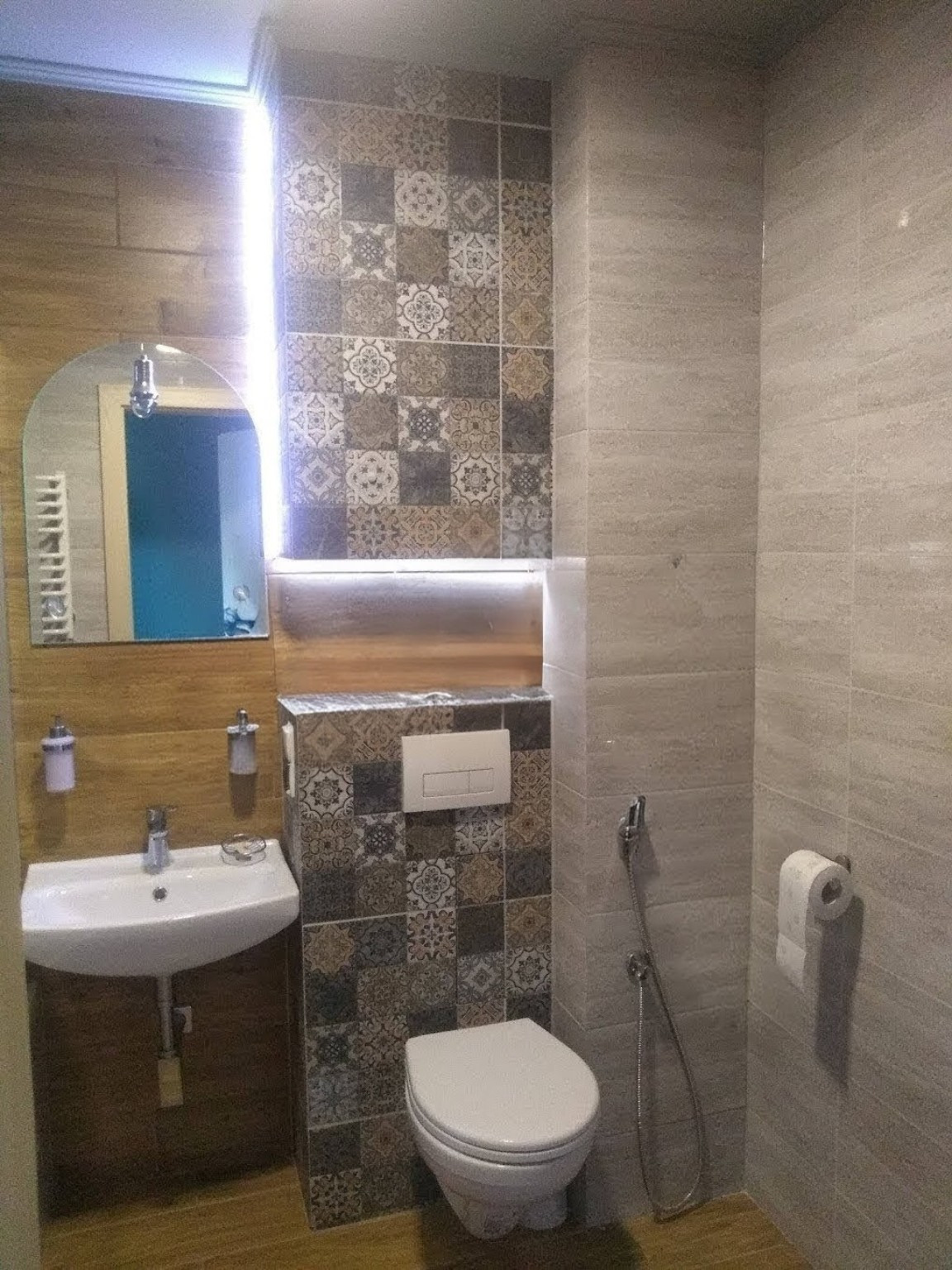 Wykończona łazienka z mozaiką na ścianie za toaletą i umywalką. Umywalka wisząca z lustrem. Na ścianach płytki imitujące drewno i kamień.