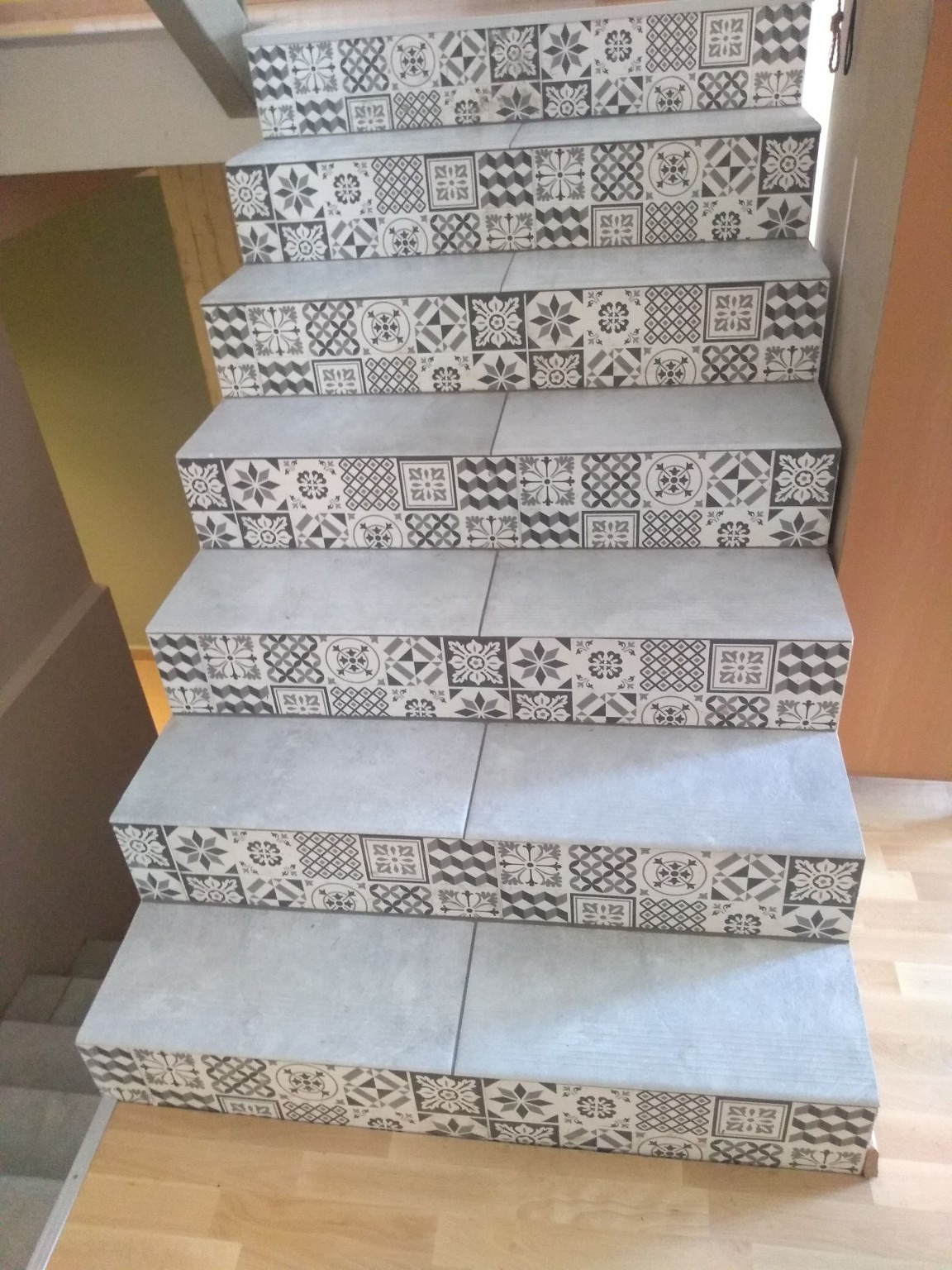 Schody wyłożone szarymi płytkami z dekoracyjnym wzorem patchworkowym na krawędziach stopni, widok z dołu, drewniana podłoga, jasne wnętrze.