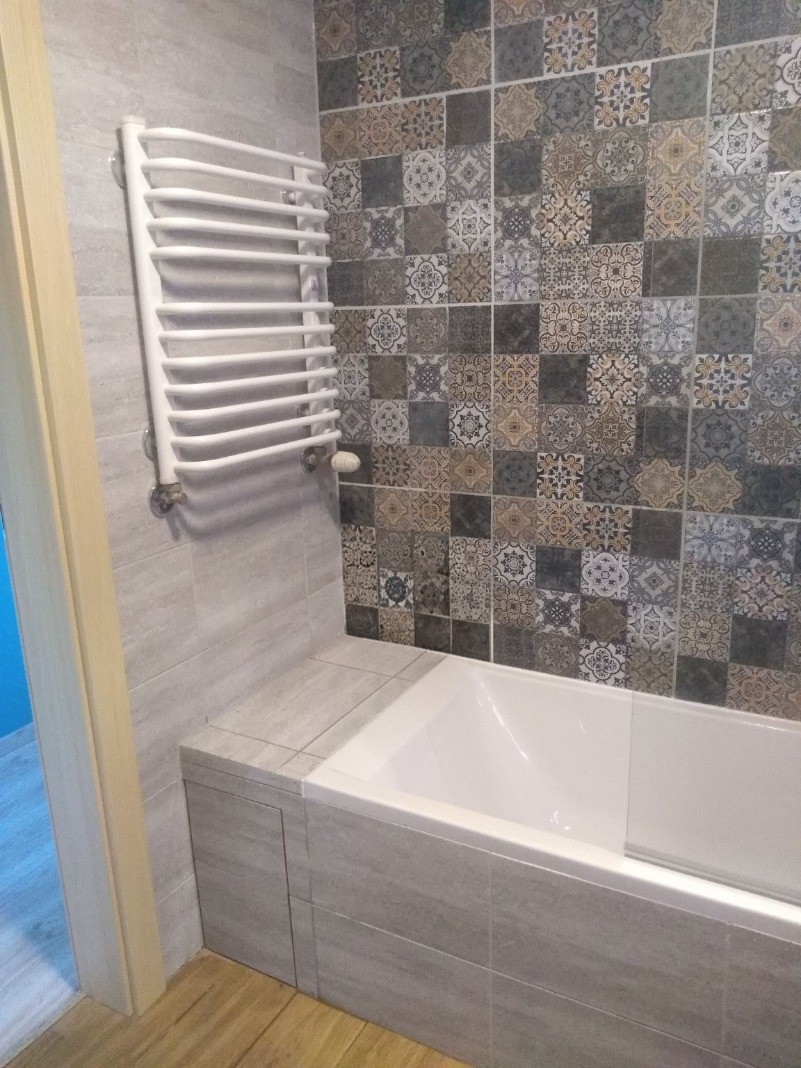 Wykończona łazienka z wanną obudowaną płytkami w odcieniach szarości i beżu. Na ścianie dekoracyjne płytki patchwork oraz biały grzejnik drabinkowy.