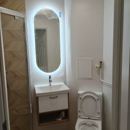 POGOTOWIE BUDOWLANE Marcin Kasiorek - Nowoczesna łazienka z podświetlanym lustrem, białą umywalką z szafką i toaletą. Widoczna kabina prysznicowa z drewnopodobnymi płytkami. Lustro owinięte folią ochronną.