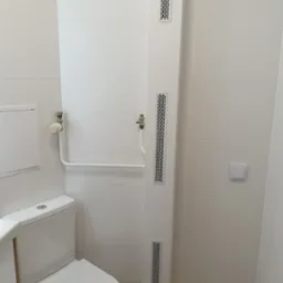 Nowoczesna łazienka w bieli: toaleta z umywalką, poziome płytki, pionowy panel z kratkami wentylacyjnymi, grzejnik i włącznik światła.