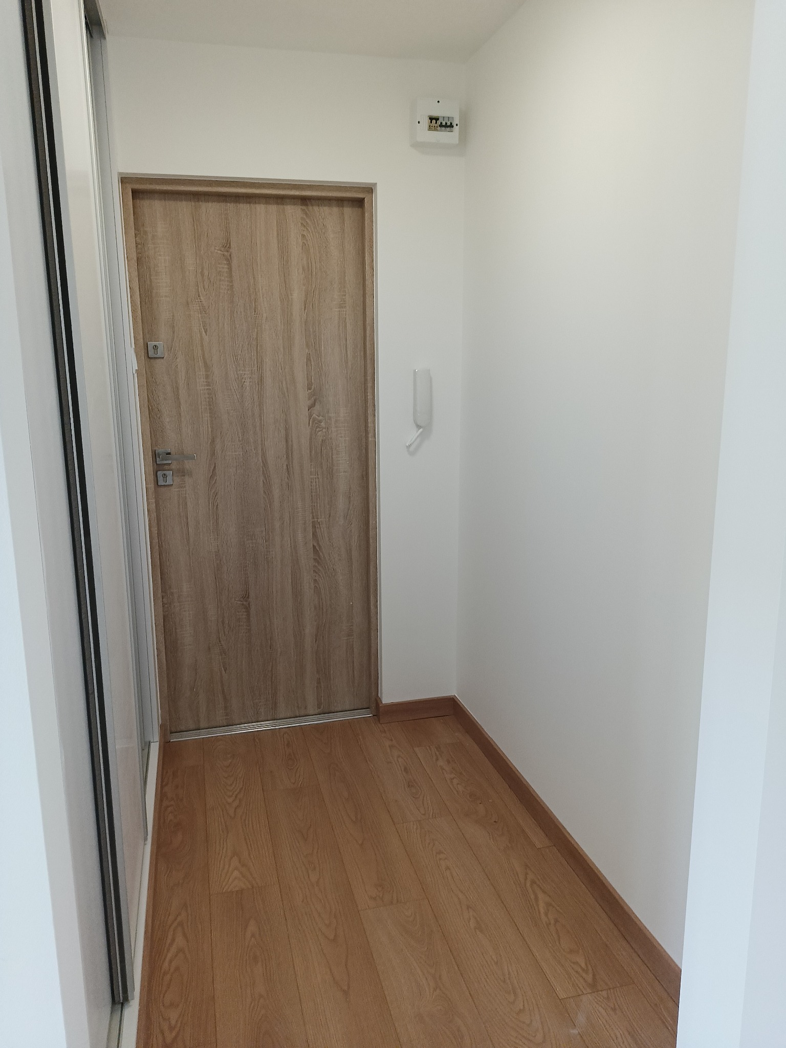 Jasne wnętrze: drewnopodobna podłoga, drzwi z klamką, białe ściany z domofonem i skrzynką elektryczną. Minimalistyczny design.