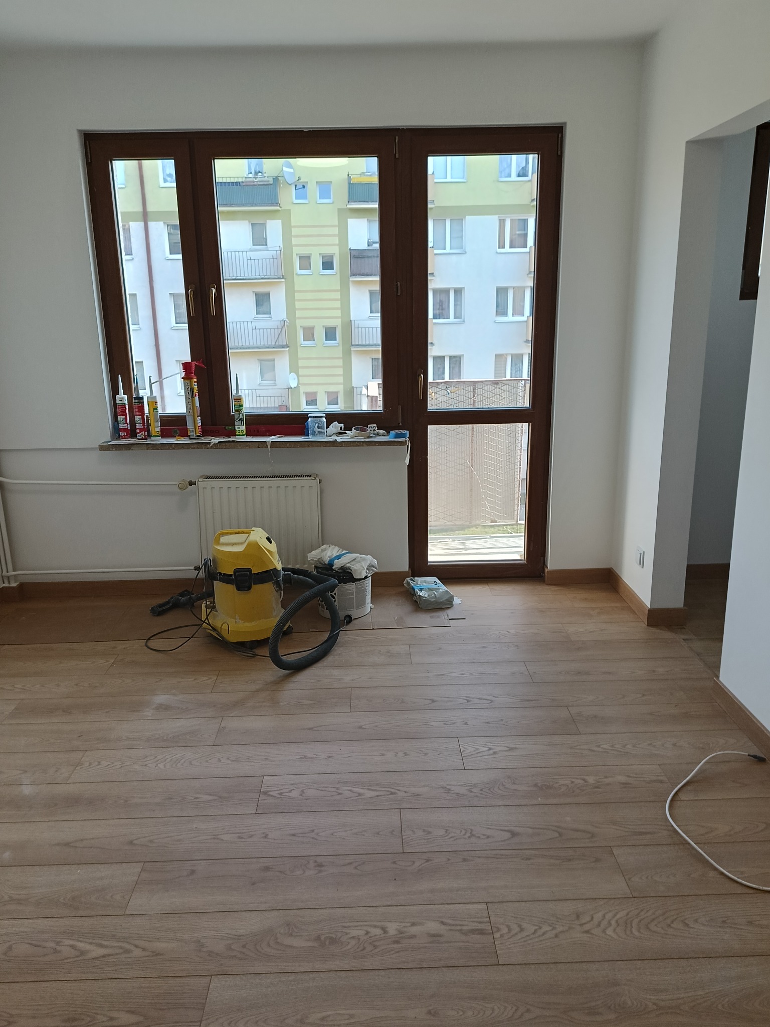Pokój po remoncie: świeżo ułożony parkiet z widocznym odkurzaczem i akcesoriami malarskimi na parapecie. W tle okno z widokiem na budynek.