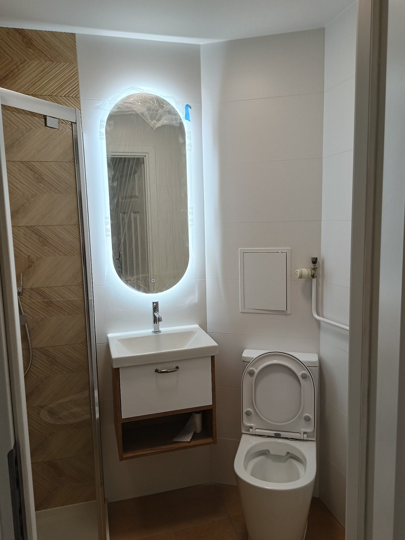 Nowoczesna łazienka z podświetlanym lustrem, białą umywalką z szafką i toaletą. Widoczna kabina prysznicowa z drewnopodobnymi płytkami. Lustro owinięte folią ochronną.