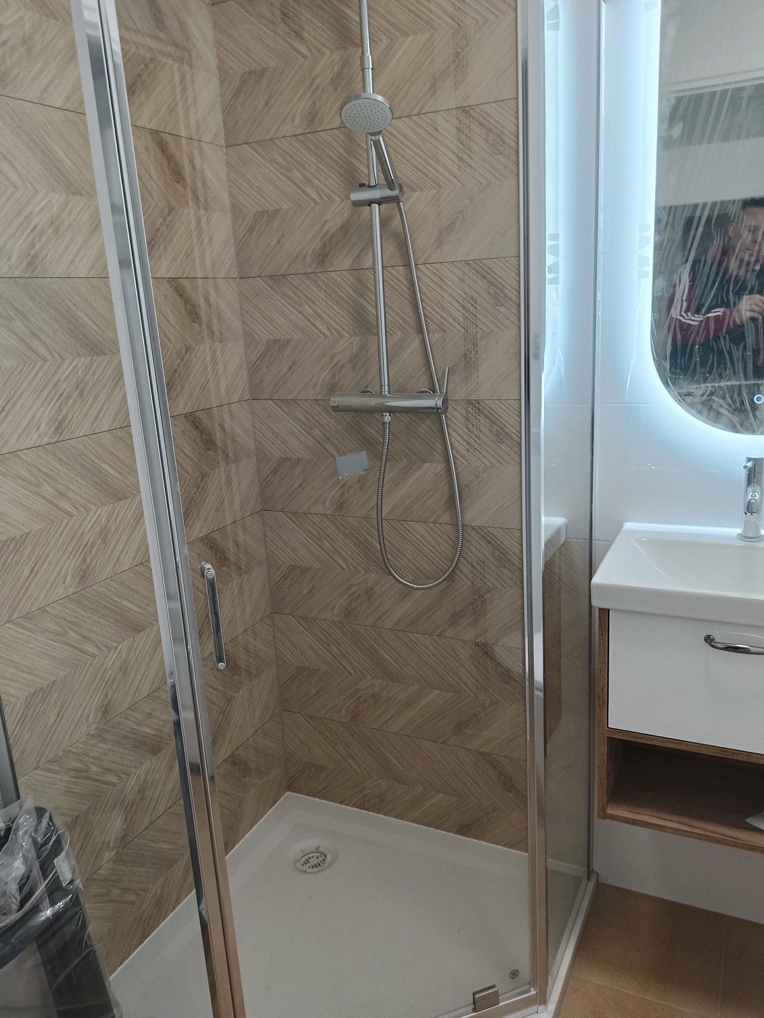 Nowoczesna kabina prysznicowa z deszczownicą i umywalką, wykończona płytkami imitującymi drewno. Elementy wciąż zabezpieczone folią ochronną.