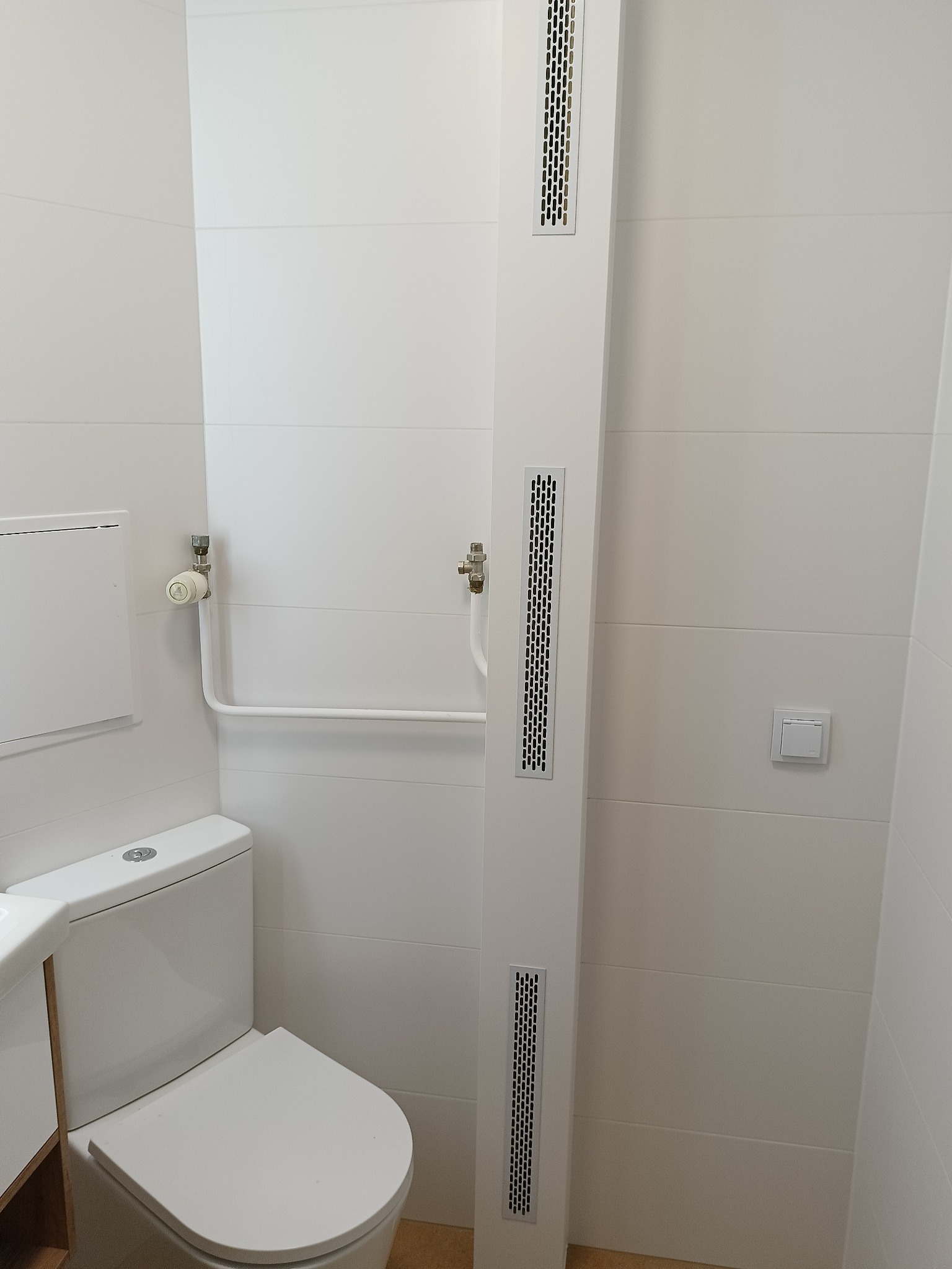 Nowoczesna łazienka w bieli: toaleta z umywalką, poziome płytki, pionowy panel z kratkami wentylacyjnymi, grzejnik i włącznik światła.