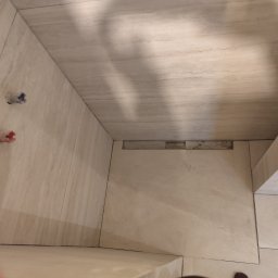 Pol-bud - Wykończona łazienka z beżowymi płytkami imitującymi marmur, widoczny odpływ liniowy i wystające rury instalacji wodnej. Minimalistyczny design.