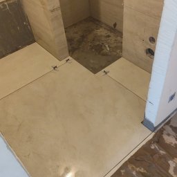 Pol-bud - Układanie beżowych płytek podłogowych w łazience, widoczne krzyżyki dystansowe i fragmenty nieukończonej posadzki w kabinie prysznicowej.