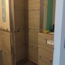 Pol-bud - Wnętrze łazienki w trakcie prac. Widoczne beżowe płytki imitujące kamień, krzyżyki dystansowe, fragment ściany z płyt gipsowo-kartonowych i narzędzia pracy.