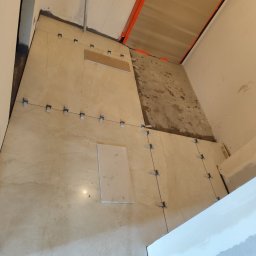 Pol-bud - Układanie beżowych płytek podłogowych z użyciem krzyżyków dystansowych. Częściowo wykończona podłoga w pomieszczeniu z widocznymi elementami instalacji.