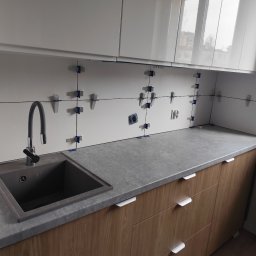 Pol-bud - Białe płytki ścienne w trakcie układania w kuchni, z widocznymi krzyżykami dystansowymi, szarą umywalką i drewnianymi szafkami. Widoczne gniazdka elektryczne.