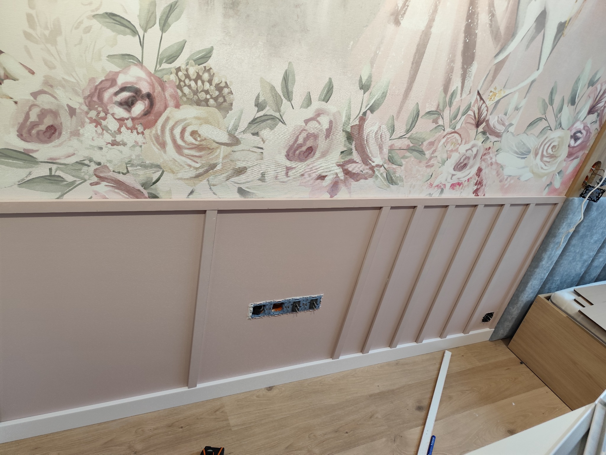 Różowa ściana z dekoracyjnym panelem i kwiatowym wzorem tapety. Widoczne gniazdka elektryczne i fragment podłogi z jasnego drewna.