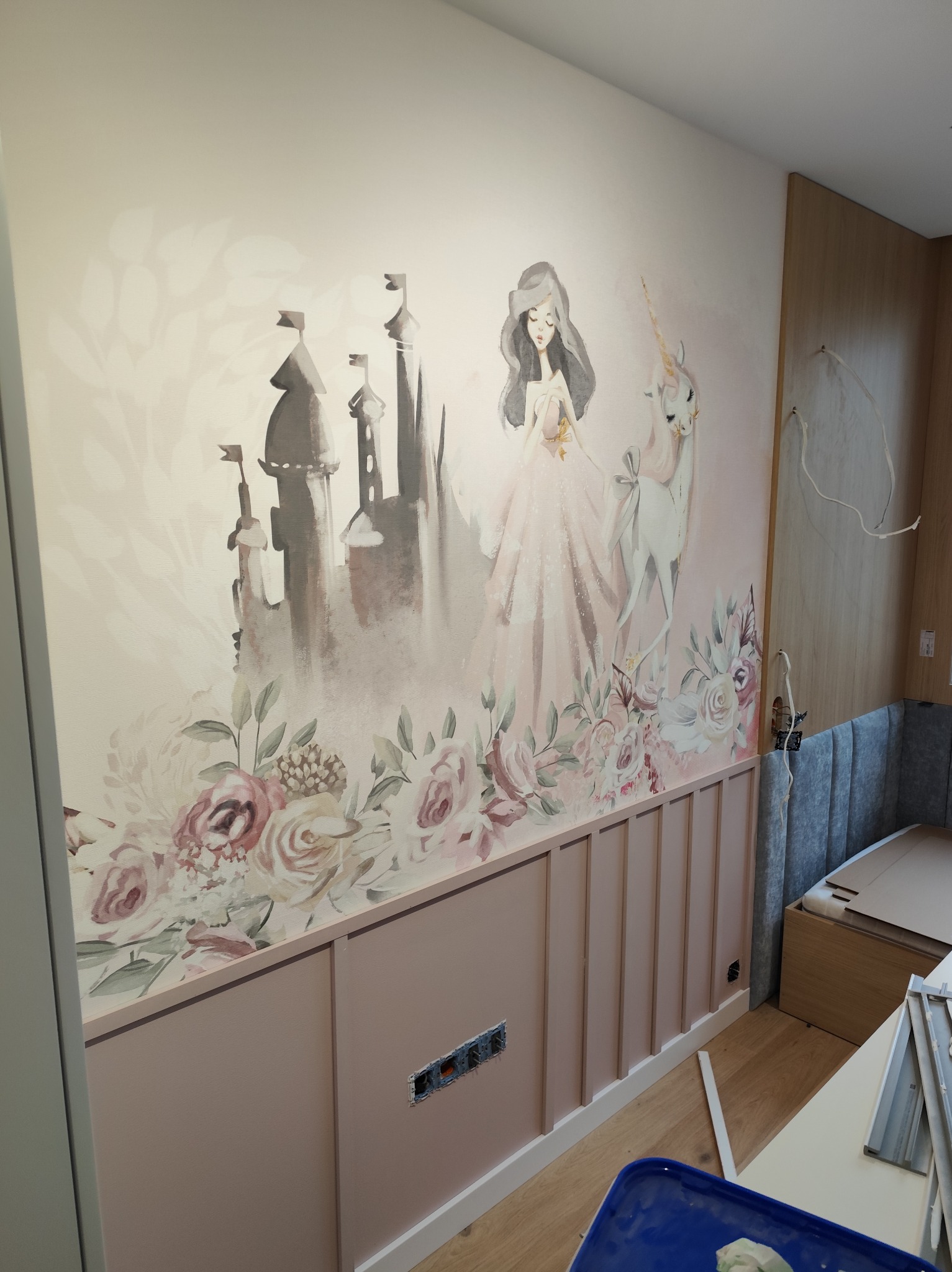 Pokój dziecięcy z dekoracyjną tapetą w pastelowych barwach, przedstawiającą księżniczkę, jednorożca i zamek. Dolna część ściany wykończona panelami w kolorze pudrowego różu.