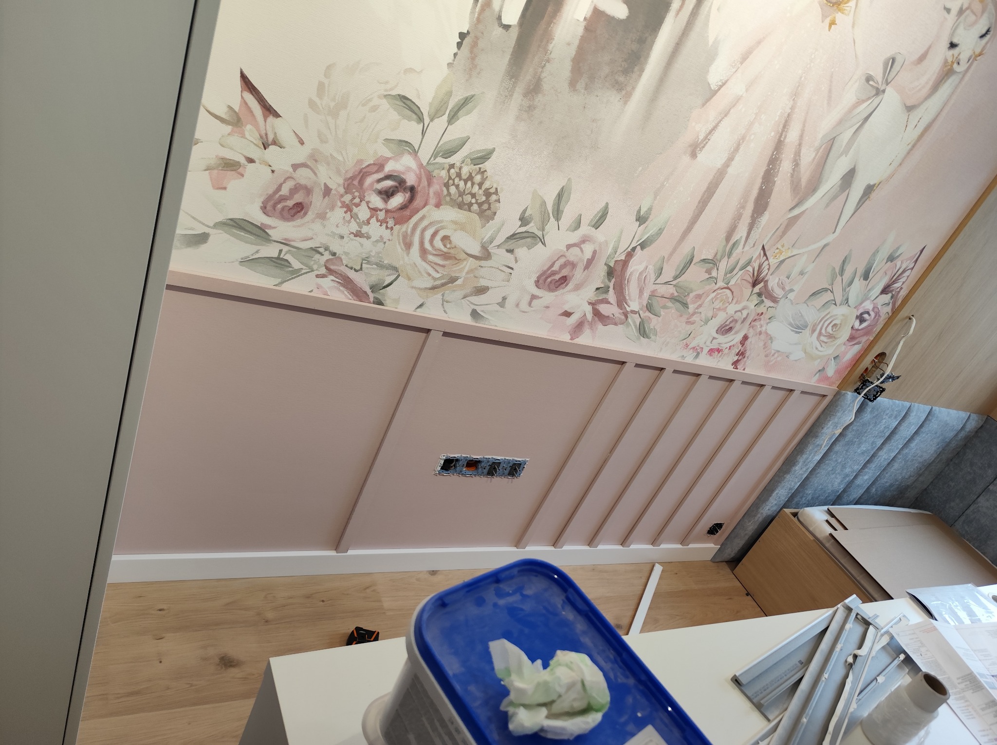 Wykończona ściana z dekoracyjnymi listwami w odcieniach różu i kwiatowym motywem na suficie. Widoczne gniazdka elektryczne i fragmenty narzędzi.