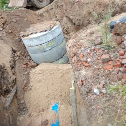 OLBUD Oliwier Brzózka - Wykop pod szambo z betonowymi kręgami, w tle koparka. Widoczne niebieskie taśmy i poziomica. Fragmenty cegieł na krawędzi wykopu. Ujęcie z góry.