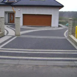 Dwukolorowa kostka brukowa ułożona w geometryczny wzór na podjeździe przed domem z brązową bramą garażową i żółtą skrzynką gazową.