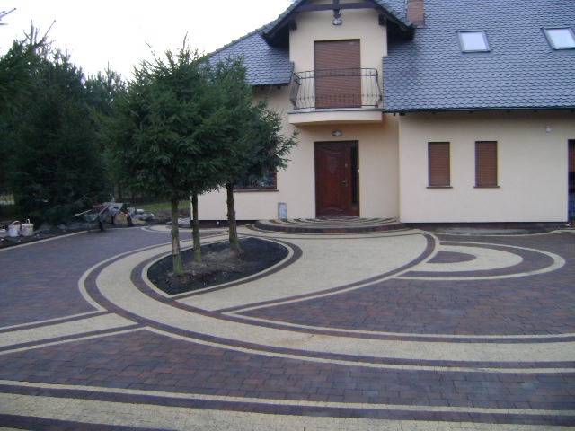 Oryginalny wzór kostki brukowej przed domem w Legnicy. Ciemnobrązowa kostka przecinana jasnymi liniami, tworząc dekoracyjny efekt. Trzy drzewka w centralnej części.