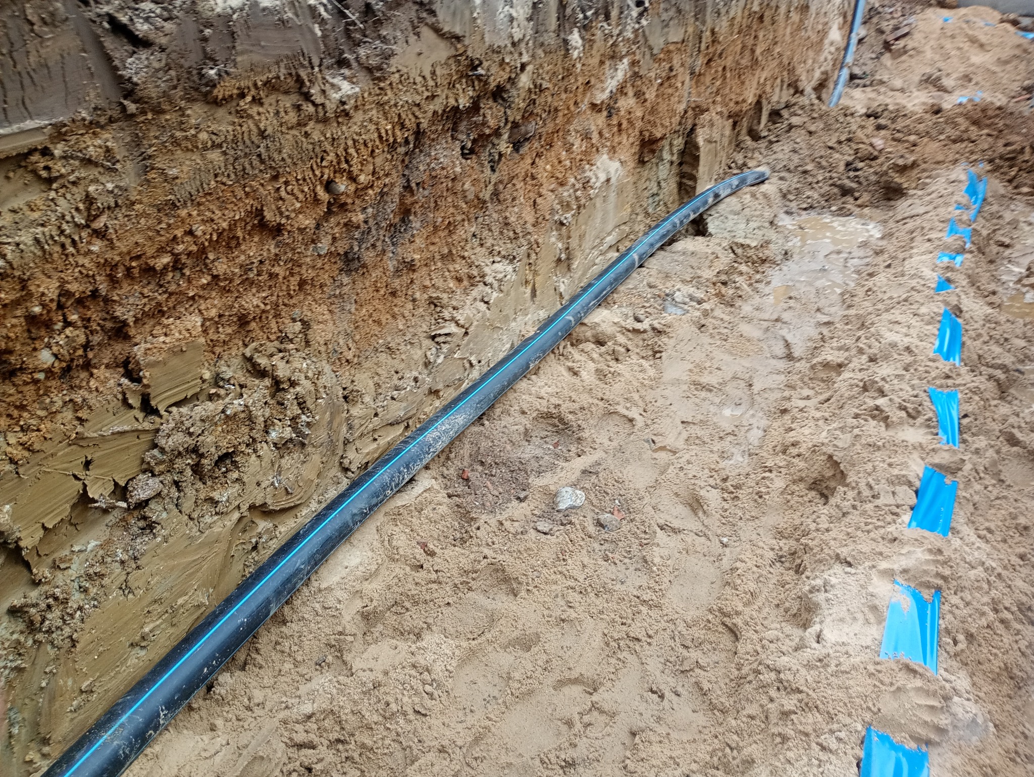 Wykop z ułożoną czarną rurą z niebieskim paskiem i niebieskimi znacznikami w piasku. Etap budowy przyłącza wodno-kanalizacyjnego.