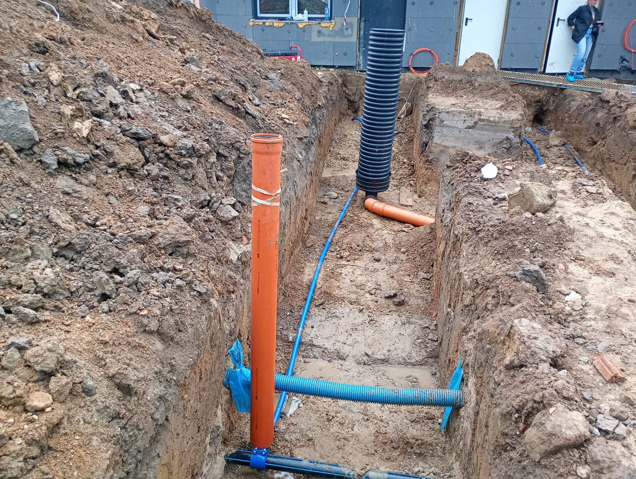 Wykop z widocznymi rurami kanalizacyjnymi w ziemi, w trakcie instalacji. Rury w kolorach pomarańczowym i niebieskim, ziemia gliniasta, fragment budynku w tle.