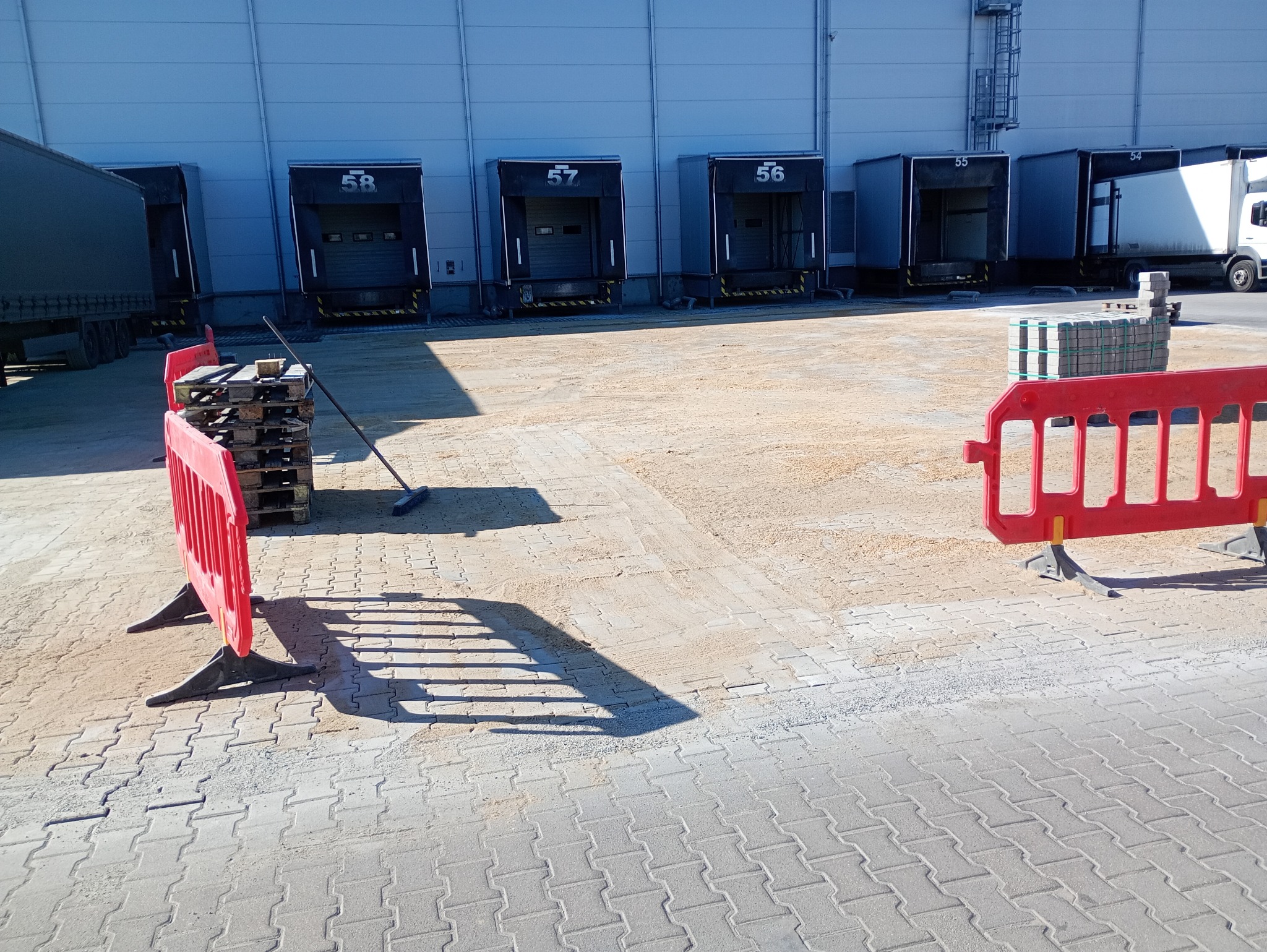 Przygotowanie terenu pod kostkę brukową: widoczne palety z kostką, bariery ochronne i fragment ułożonej nawierzchni w tle magazynów.