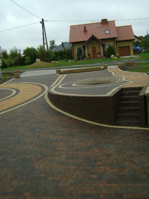 Wzorzysta nawierzchnia z kostki brukowej w odcieniach brązu i beżu, prowadząca do domu z czerwonym dachem. Schody z cegły i elementy małej architektury.