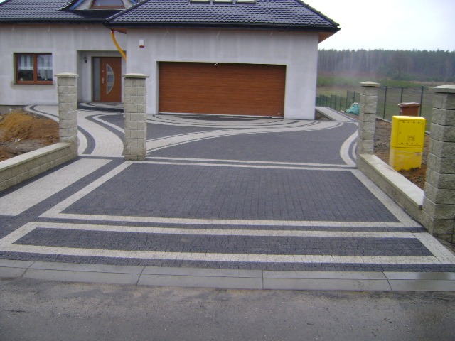 Dwukolorowa kostka brukowa ułożona w geometryczny wzór na podjeździe przed domem z brązową bramą garażową i żółtą skrzynką gazową.