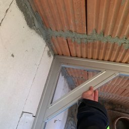 Golon Expert sp. z o.o. - Montaż metalowej ramy w narożniku pomieszczenia z białymi bloczkami i ceglanym stropem. Widoczna ręka przytrzymująca element konstrukcji.