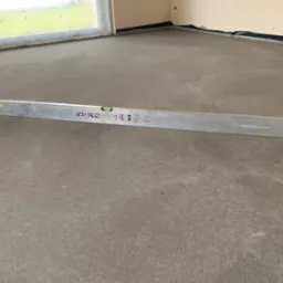 Wyrównana, świeża wylewka betonowa podłogi w pomieszczeniu z oknem, sprawdzana poziomicą. Widoczne zabezpieczenia ścian folią. Szary beton, srebrna poziomica.