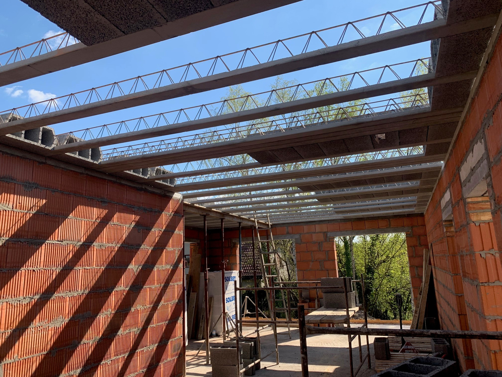 Wnętrze budynku w trakcie budowy z czerwonej cegły. Widoczne zbrojenie stropu i fragment nieba przez otwory. Ceglana ściana w cieniu rzuca prążkowane cienie.