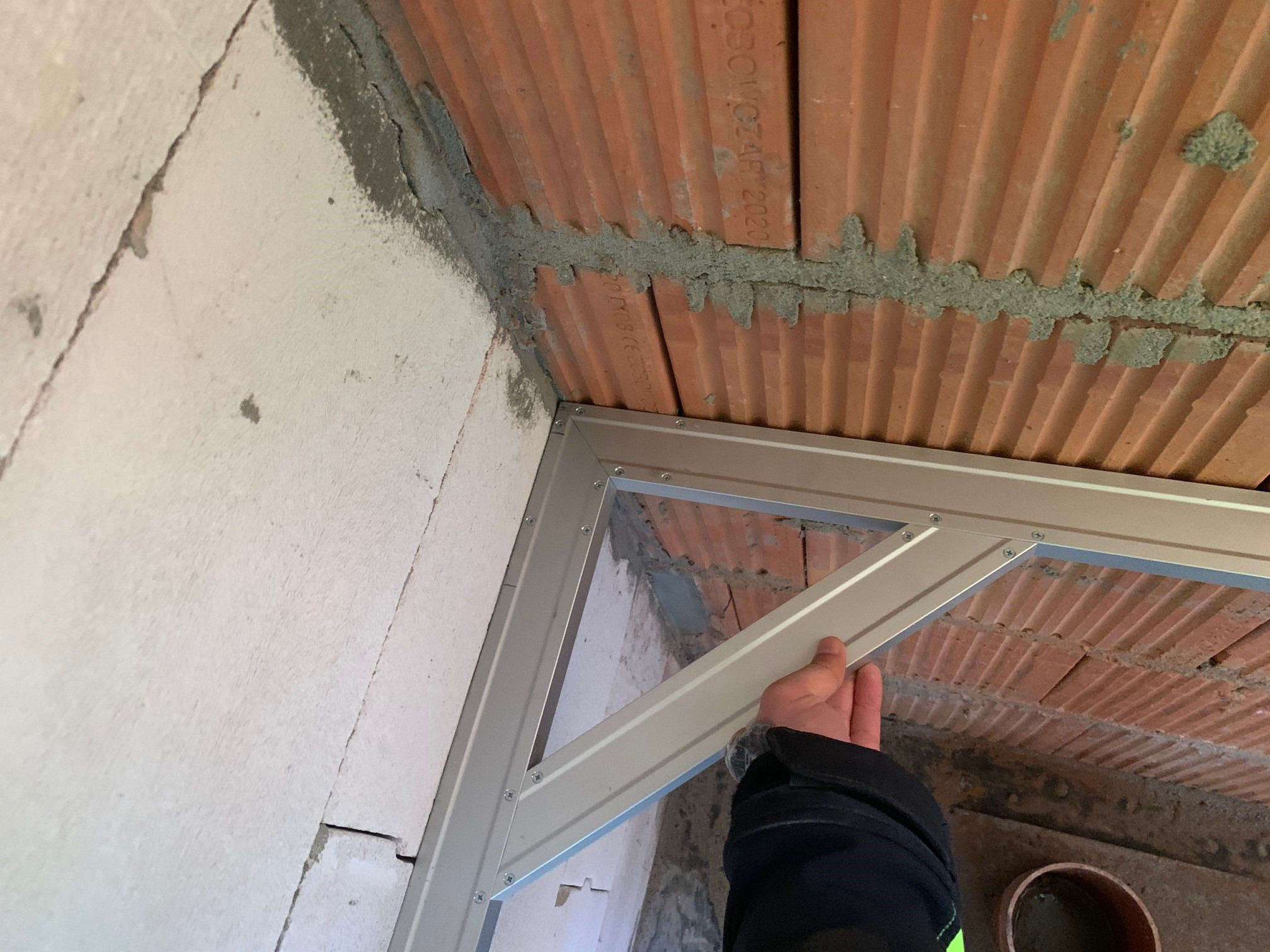 Montaż metalowej ramy w narożniku pomieszczenia z białymi bloczkami i ceglanym stropem. Widoczna ręka przytrzymująca element konstrukcji.